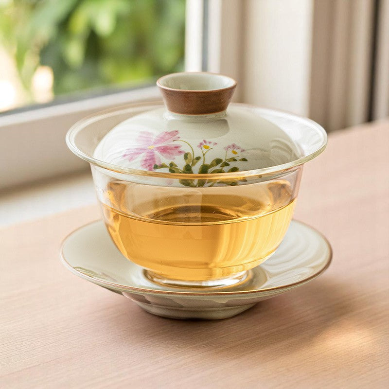 Gaiwan with floral design on a saucer, placed on a wooden surface with a window in the background.