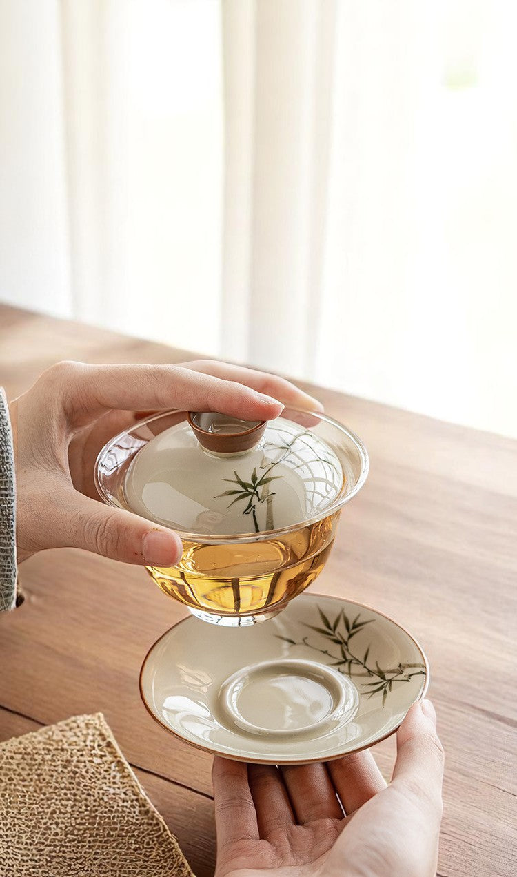 Person holding a glass gaiwan with a ceramic lid on a wooden surface