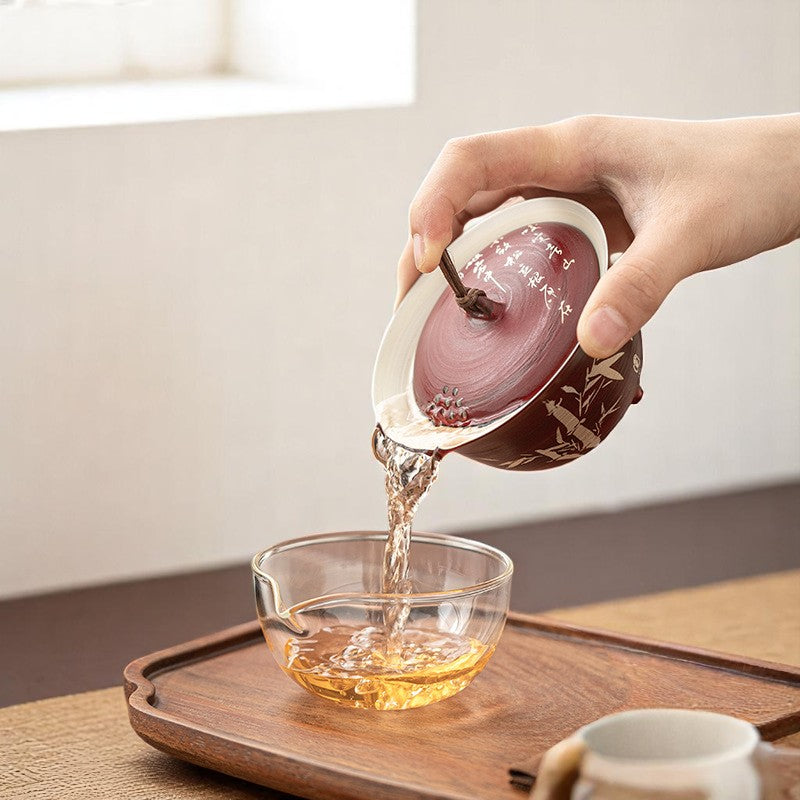 Person pouring tea from a ceramic teapot into a glass bowl on a wooden tray.