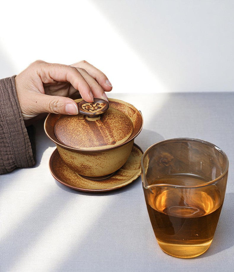 Person holding a ceramic tea infuser over a teacup with a glass of tea on a light surface.