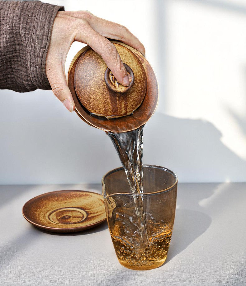 Person pouring tea from a stoneware gaiwan into a glass on a light surface.